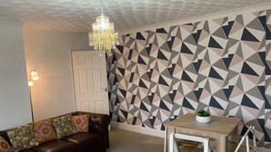 Ein Wohnzimmer mit einem braunen Ledersofa, einem weißen Couchtisch mit Pflanze, einem Holzesstisch mit Stühlen, einer Wand mit geometrischem Muster und einer Kronleuchter.