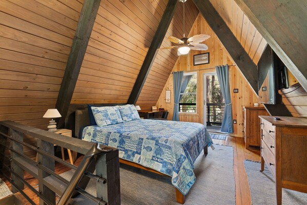 A bedroom with a wooden ceiling, a bed with a blue patterned bedspread, a desk, and a window with curtains. - Fox Den~Perfect Escape for Birders (Dauphin Island)