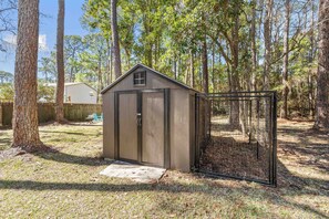 A small, single-story shed with a metal door and window, surrounded by trees and a grassy yard. - Fox Den~Perfect Escape for Birders (Dauphin Island)