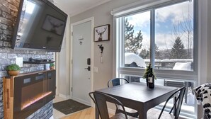 A modern kitchen with a TV, a fireplace, a dining area with a table and chairs, and a large window with a view of snow-covered trees. - Cozy Chalet in the Mont Tremblant Ski Village (Mont-Tremblant)