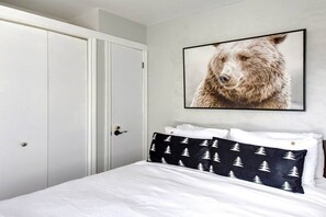 A bedroom with a white bed, a black and white patterned pillow, a framed bear picture on the wall, and a white wardrobe.
