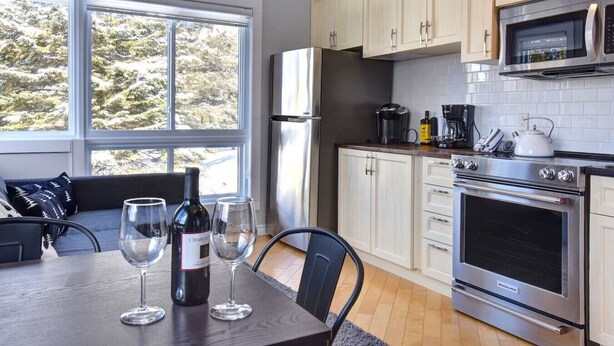 A modern kitchen with stainless steel appliances, a dining area with a table set for two, and a large window offering a view of the outdoors. - Cozy Chalet in the Mont Tremblant Ski Village (Mont-Tremblant)