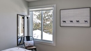 A bedroom with a bed, a chair, a window with a view of trees, and a framed picture on the wall. - Cozy Chalet in the Mont Tremblant Ski Village (Mont-Tremblant)