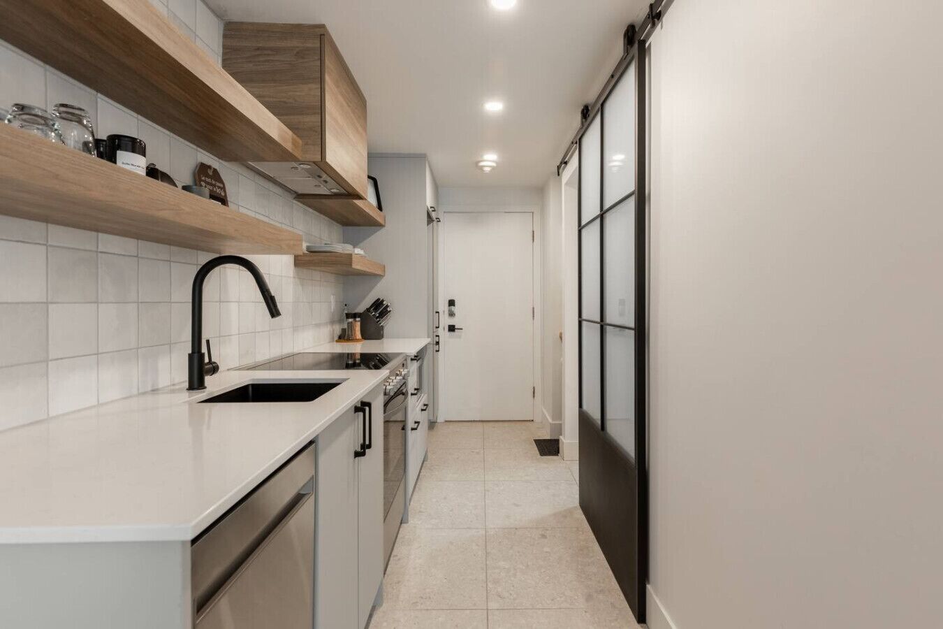 A narrow kitchen with white tiles, a black faucet, wooden shelves, and a sliding door.