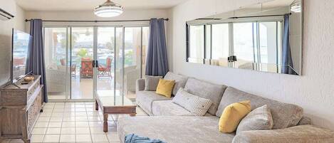 A living room with a sofa, a coffee table, and a TV. There is a sliding glass door leading to an outdoor area with a pool.