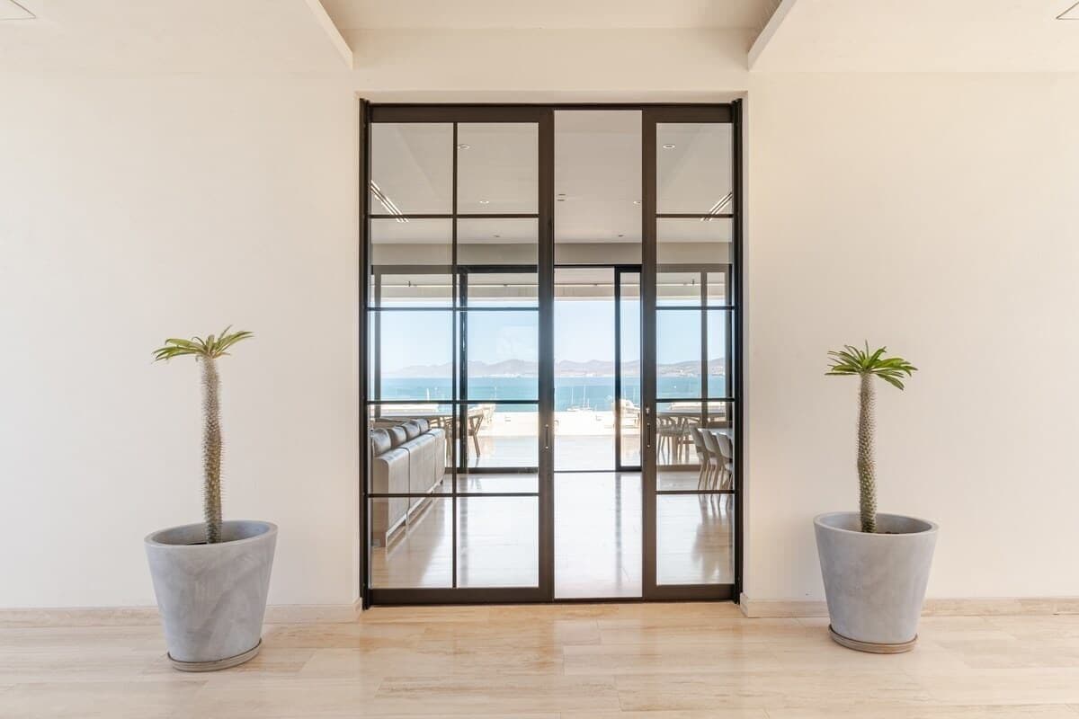 A modern interior with a sliding glass door leading to a balcony with a view of the sea. There are two potted plants on either side of the door.