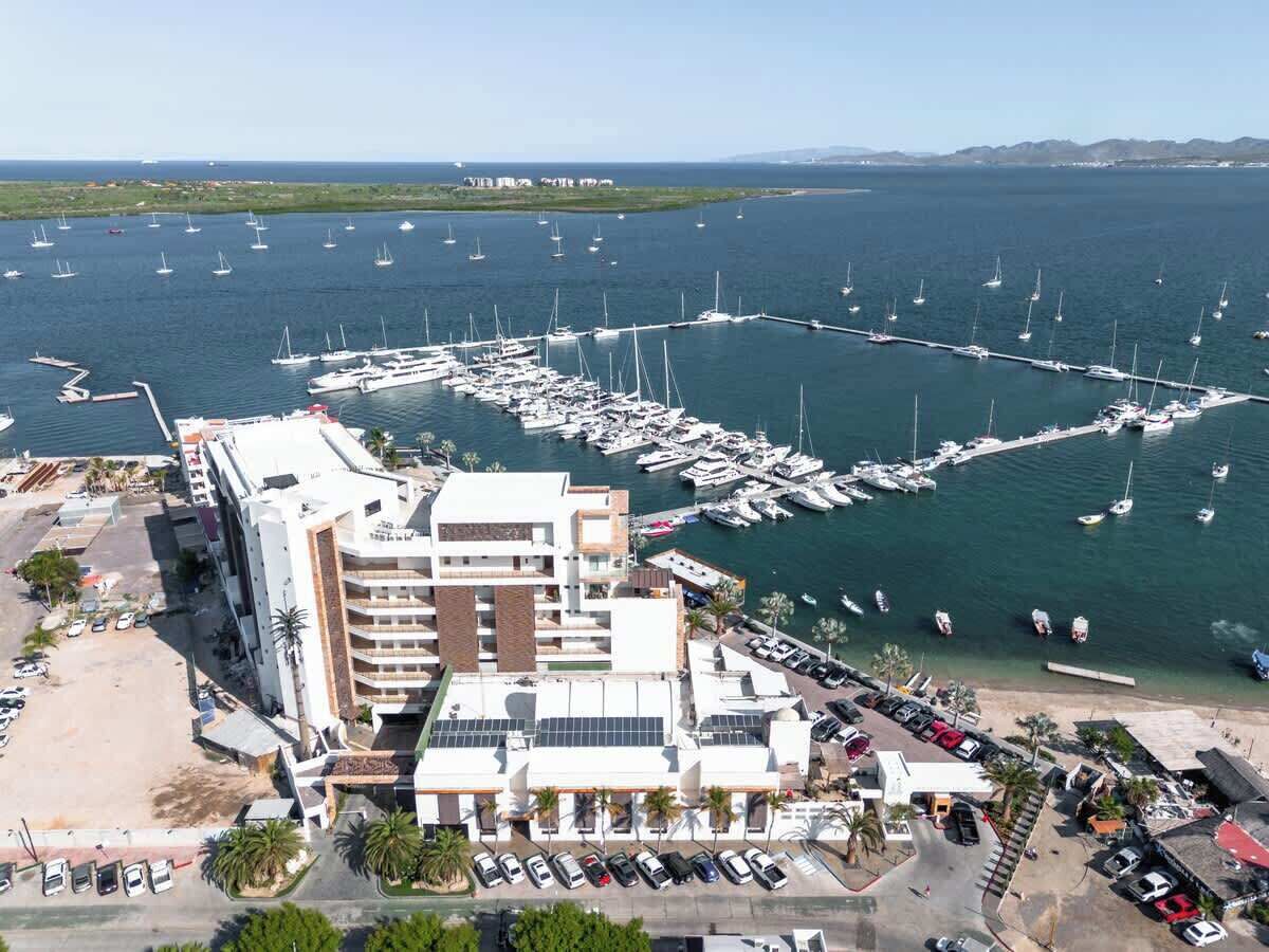 Un puerto deportivo con numerosas embarcaciones amarradas, un edificio moderno de varios pisos y un estacionamiento con varios automóviles.