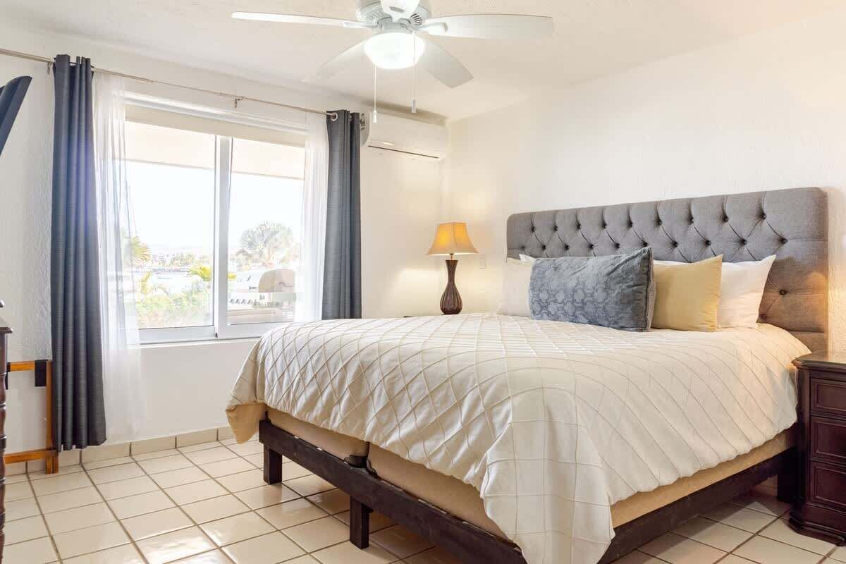 Un dormitorio con cama, ventilador de techo, ventana con cortinas y mesita de noche con lámpara.