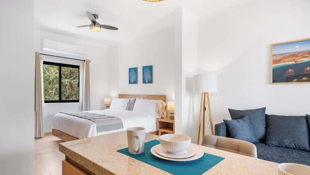 A modern hotel room with a bed, a sitting area with a sofa, a small kitchen area with a counter and a bowl, and a ceiling fan. - Pineda Suite 202 with Parking (La Paz)