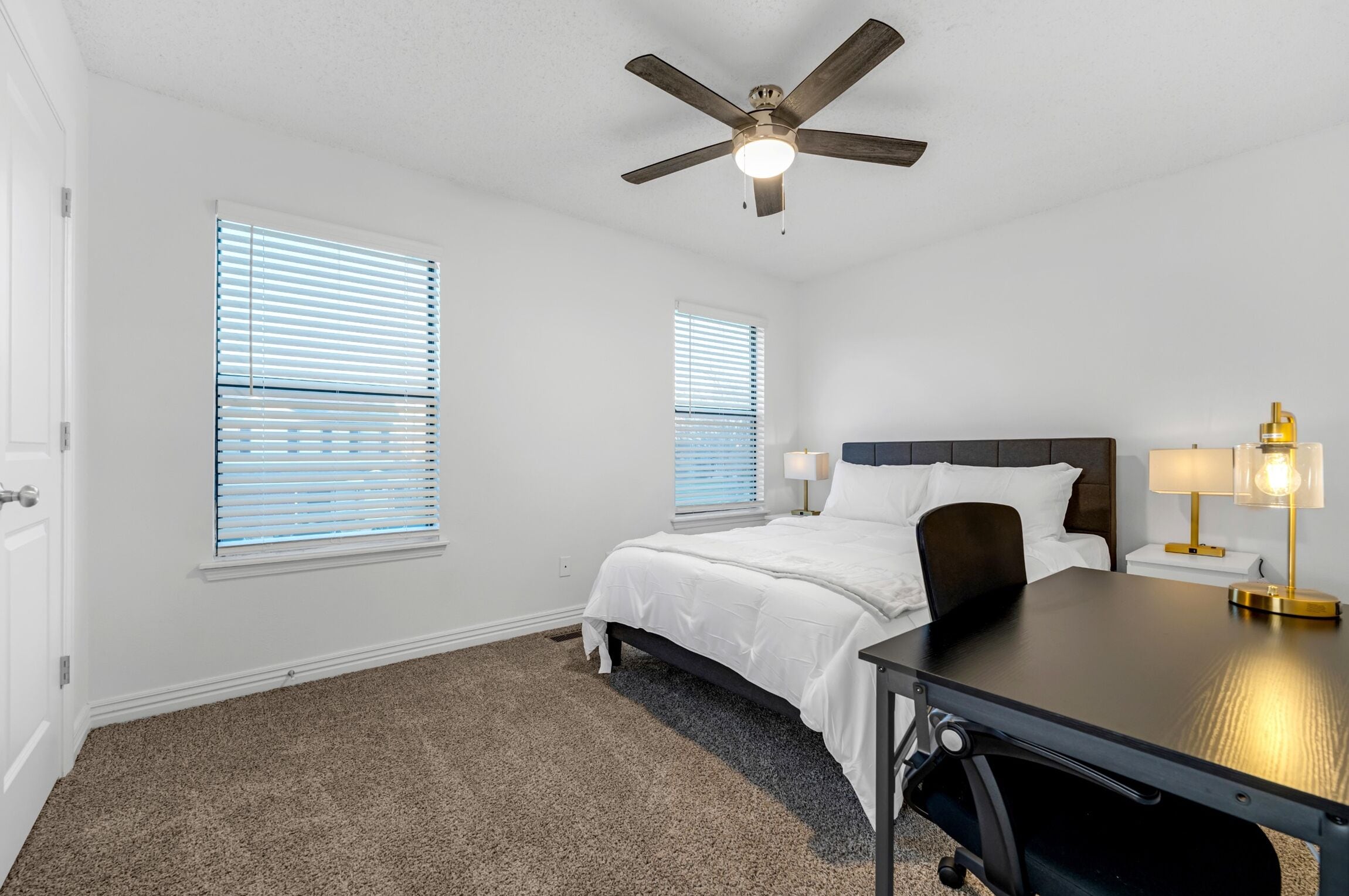 A bedroom with a bed, a desk, two bedside tables with lamps, a ceiling fan, and two windows with blinds.