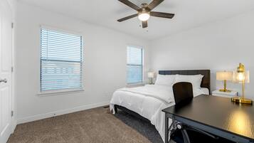 A bedroom with a bed, a desk, two bedside tables with lamps, a ceiling fan, and two windows with blinds.