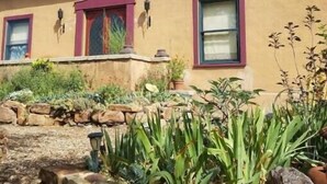 A two-story house with a stone foundation, a metal roof, and a garden with various plants and a stone wall. - Romantic Adobe Casita, Hot tub, Kitchen, Woodstove, astounding views, historic. (Los Cerrillos)