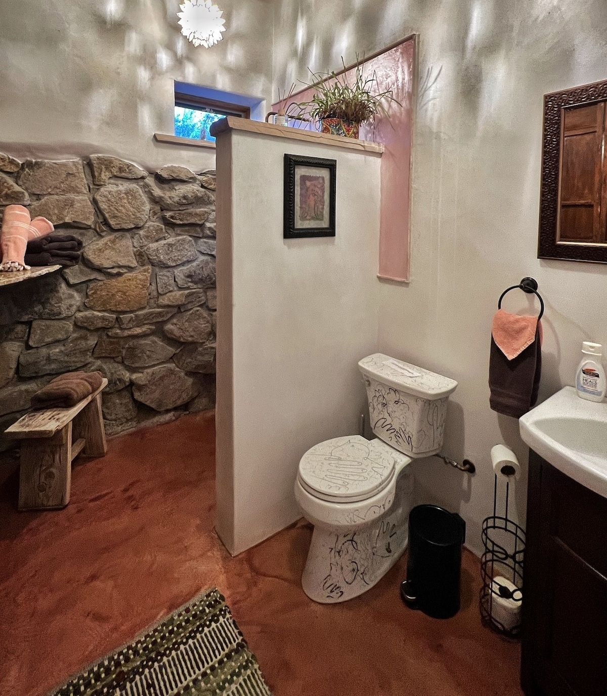 Une salle de bain avec un mur en pierre, des toilettes ornées de motifs floraux, un lavabo et une petite étagère avec des serviettes.