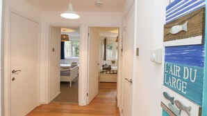 A hallway with wooden flooring, white walls, and a door leading to a bedroom with a bed and a window. - Sea House (Littlehampton)