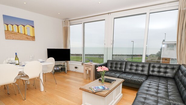 A modern living room with a large black leather sofa, a white dining table with chairs, a flat-screen TV, and a painting of beach huts on the wall. - Sea House (Littlehampton)