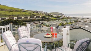 A terrace with outdoor seating and a table set with food and drinks, overlooking a coastal village with houses and a hill in the background.
