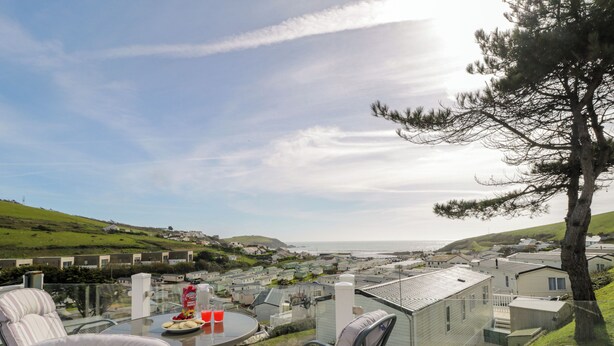 A patio with outdoor seating, a table with food and drinks, and a view of a coastal village with houses and hills. - Ocean Pearl (Kingsbridge)