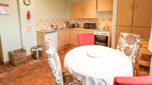 A compact kitchen with wooden cabinets, a stove, a microwave, and a sink. There is a dining table with a white tablecloth and a red chair. - Ratty's Retreat (Spilsby)
