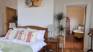 Un dormitorio con cama de madera, mesitas de noche, una lámpara, una planta en maceta y un baño visible a través de una puerta abierta.