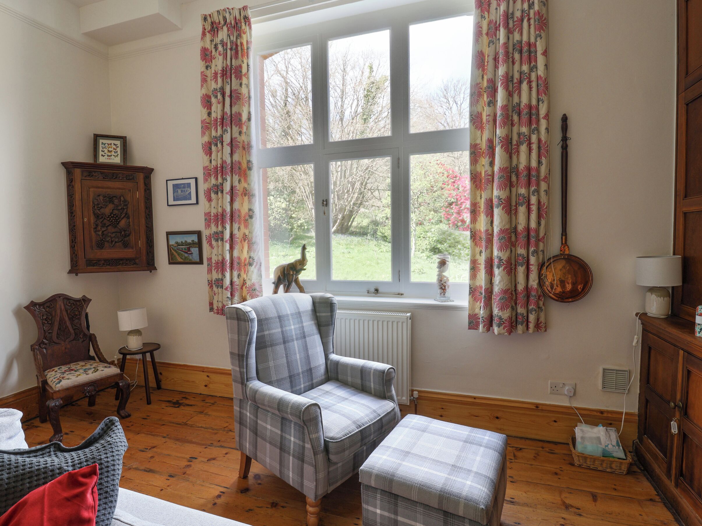 A cozy room with a plaid armchair, a matching ottoman, a wooden floor, a window with floral curtains, a radiator, and a wall-mounted lamp.