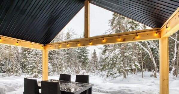 Un espacio de estar al aire libre cubierto, con una mesa y sillas, rodeado de árboles cubiertos de nieve.