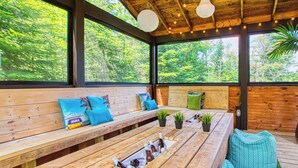An outdoor wooden deck with benches and a table, surrounded by large windows offering a view of greenery. - Alpha-O | Spa | Charlevoix par Alchalet (Petite-Rivière-Saint-François)
