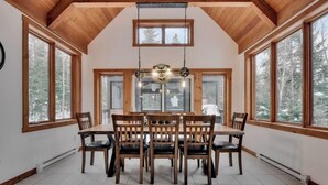 A dining area with a wooden table and chairs, large windows, and a hanging light fixture. - Alpha-O | Spa | Charlevoix par Alchalet (Petite-Rivière-Saint-François)