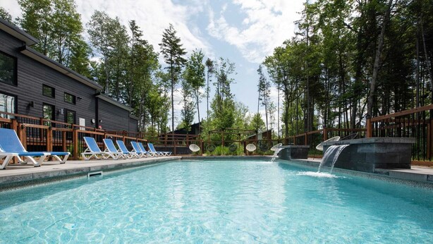 A swimming pool with lounge chairs, a waterfall feature, and a building with a dark facade. - Apollon - Piscine, Spa, Sauna by Alchalet (Pont-Rouge)
