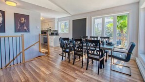 A modern kitchen with white cabinets, a dining area with a wooden table and chairs, and large windows with a view of greenery. - La Petite Ourse Cozy Getaway in Charlevoix  Spa (Petite-Rivière-Saint-François)