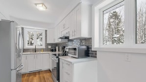 A modern kitchen with white cabinets, a microwave, and a window with a snowy view. - La Petite Ourse Cozy Getaway in Charlevoix  Spa (Petite-Rivière-Saint-François)