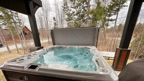 A hot tub with a wooden exterior, a bench, and a view of trees and a house in the background. - Chalet charmant Vacances avec Spa by Alchalet (Scott)