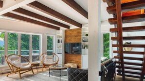 A spacious living room with large windows, a wooden ceiling, a black leather sofa, wicker chairs, and a glass coffee table.