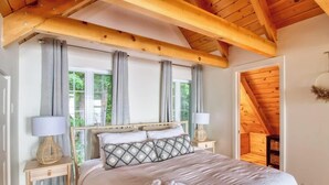 A bedroom with a wooden ceiling, a bed with a patterned headboard, two bedside lamps, and a view of greenery through the window.