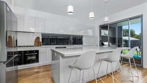 Une cuisine moderne avec des meubles blancs, un îlot central doté de tabourets de bar et une grande porte-fenêtre coulissante donnant sur un espace extérieur.