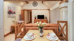 A dining area with a wooden table set for two, a vase with flowers, and a television in the background.
