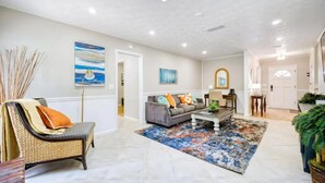 A spacious living room with a grey sofa, a coffee table, and a patterned rug. There is a chair with a cushion, a lamp, and a painting on the wall.