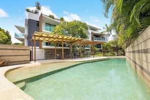 A modern pool area with a clear pool, a wooden pergola, and a multi-story building with balconies. - Peregian on the Beach Apt 1 (Peregian Beach)