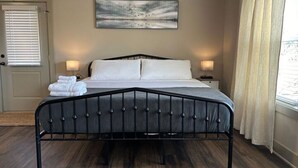 A bedroom with a bed, two bedside lamps, a door with blinds, a window with curtains, and a painting on the wall. - Choctaw Ridge King Bed-Private Deck-Gas Firepit (Dahlonega)