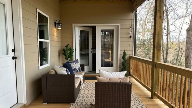 A covered porch with two wicker sofas and a matching ottoman, a wooden deck, and a view of trees and a building in the background. - Choctaw Ridge King Bed-Private Deck-Gas Firepit (Dahlonega)