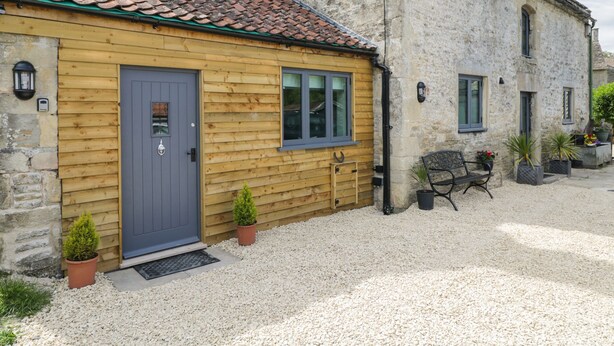 A rustic building with a wooden facade, a grey door, and a small porch area with potted plants and a bench. - Chequers Barn (Corsham)