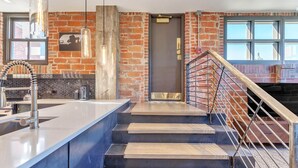 A modern kitchen with a large island, a brick wall, and a staircase with metal railings.