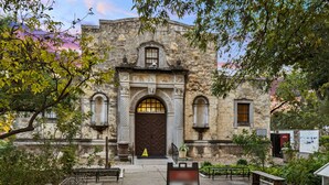 A historic building with a large arched entrance, a stone facade, and a small garden area with benches and a signboard.