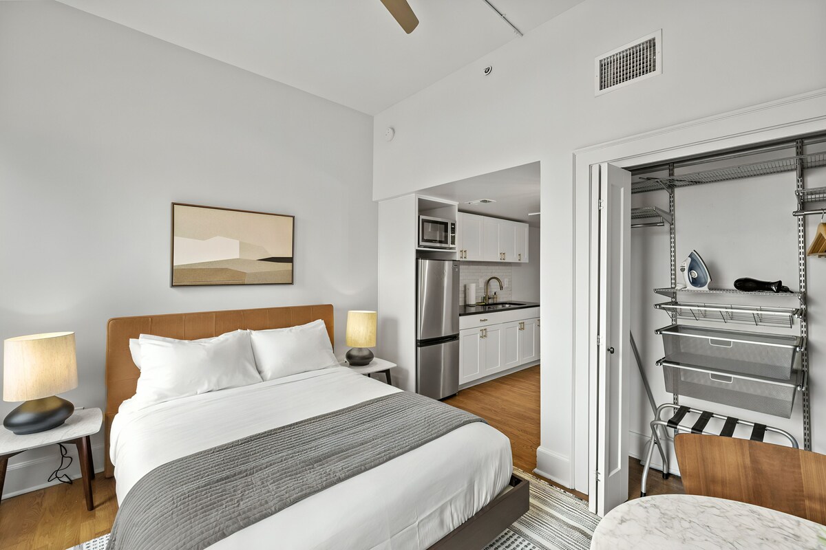 A modern hotel room with a large bed, a bedside table with lamps, a view of the kitchen, and a walk-in closet with shelves.