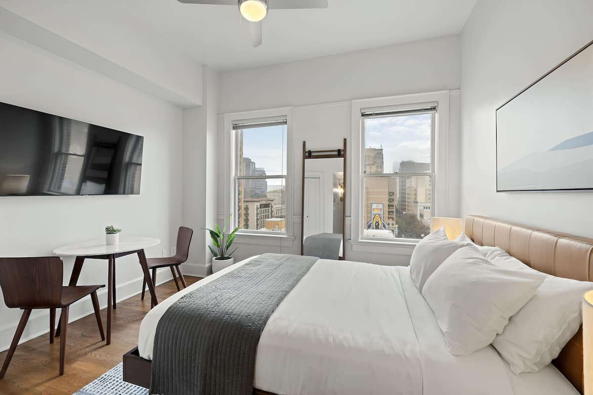 Una habitación de hotel moderna con cama, escritorio y silla. Hay un televisor montado en la pared y una ventana con vistas a la ciudad.
