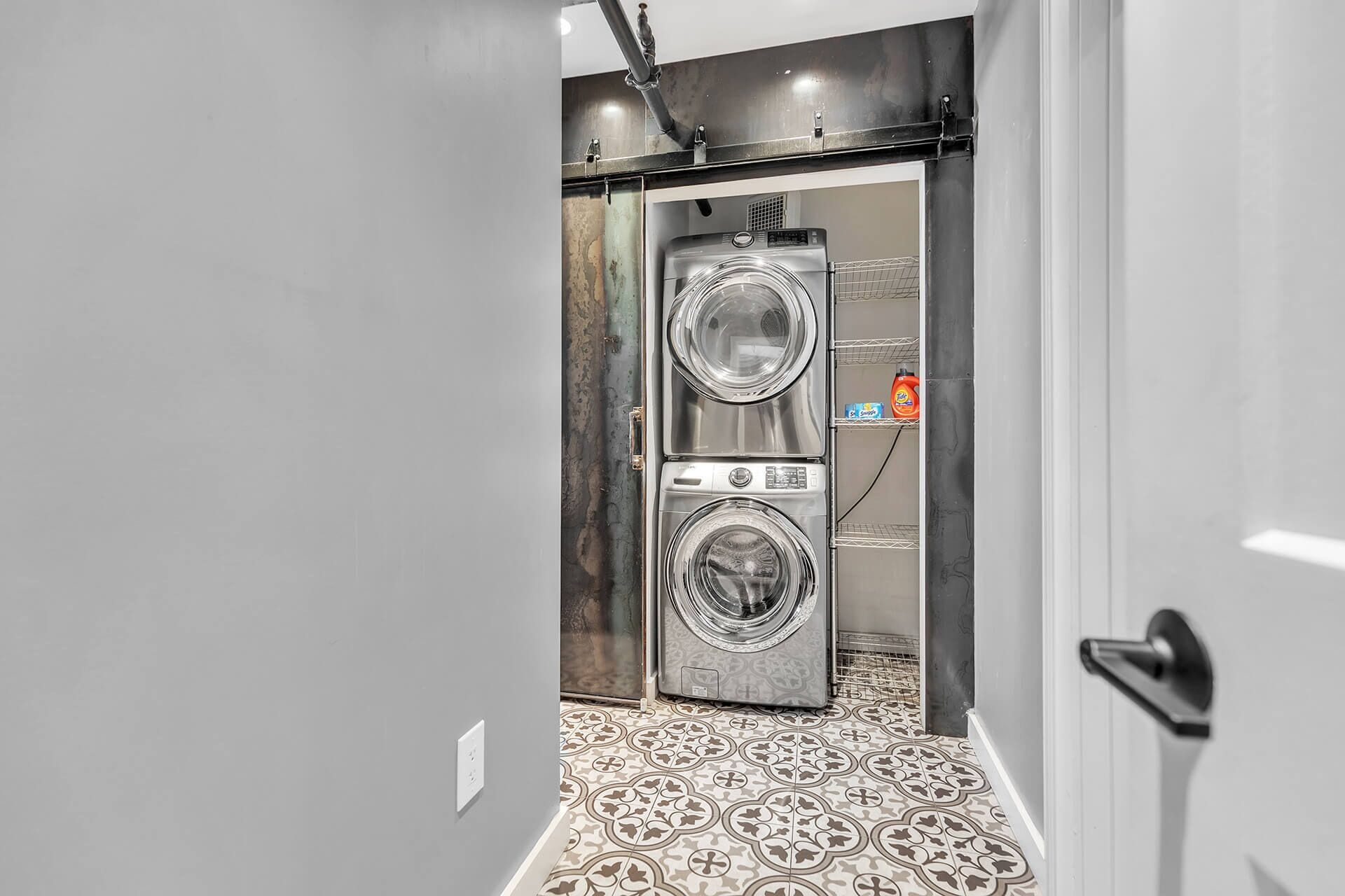 A compact, modern laundry room with two stacked front-loading washing machines, a small shelf with detergent bottles, and patterned floor tiles.