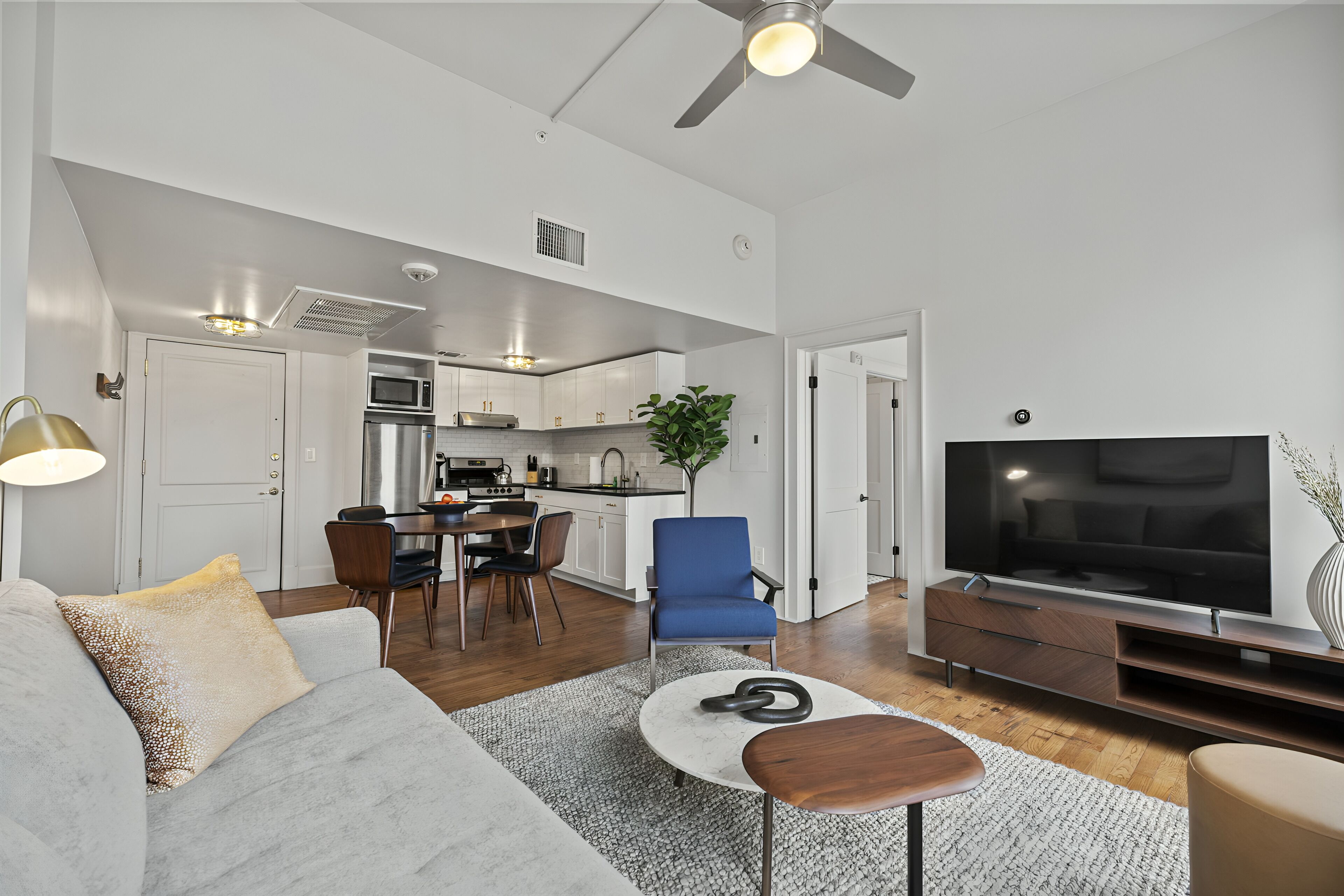 A modern living room with a sofa, coffee table, and TV. A kitchen with appliances and a dining area is visible in the background.