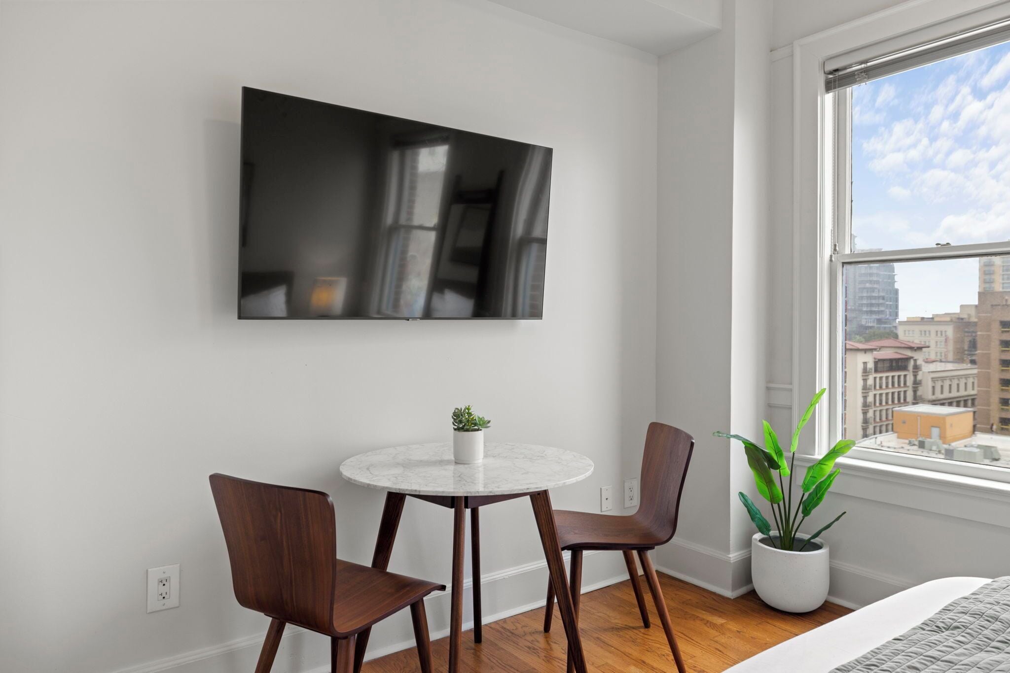 A modern room with a round table, two chairs, a TV mounted on the wall, and a large window with a city view.