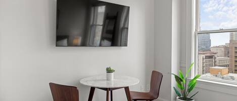 A modern room with a round table, two chairs, a TV mounted on the wall, and a large window with a city view.