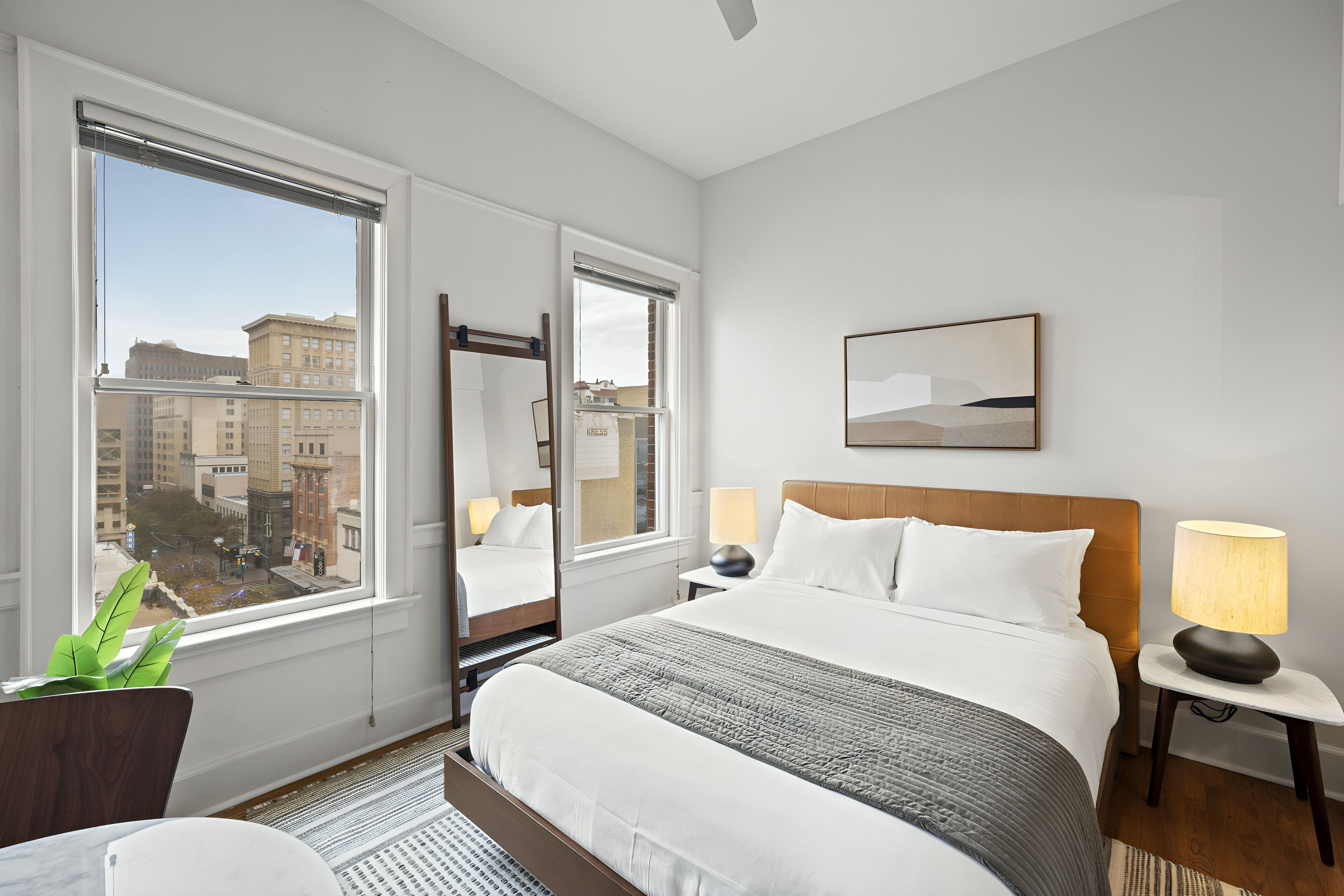 A hotel room with a large window, a bed, a bedside table with a lamp, and a mirror.