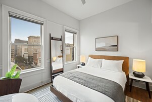 A hotel room with a large window, a bed, a bedside table with a lamp, and a mirror.
